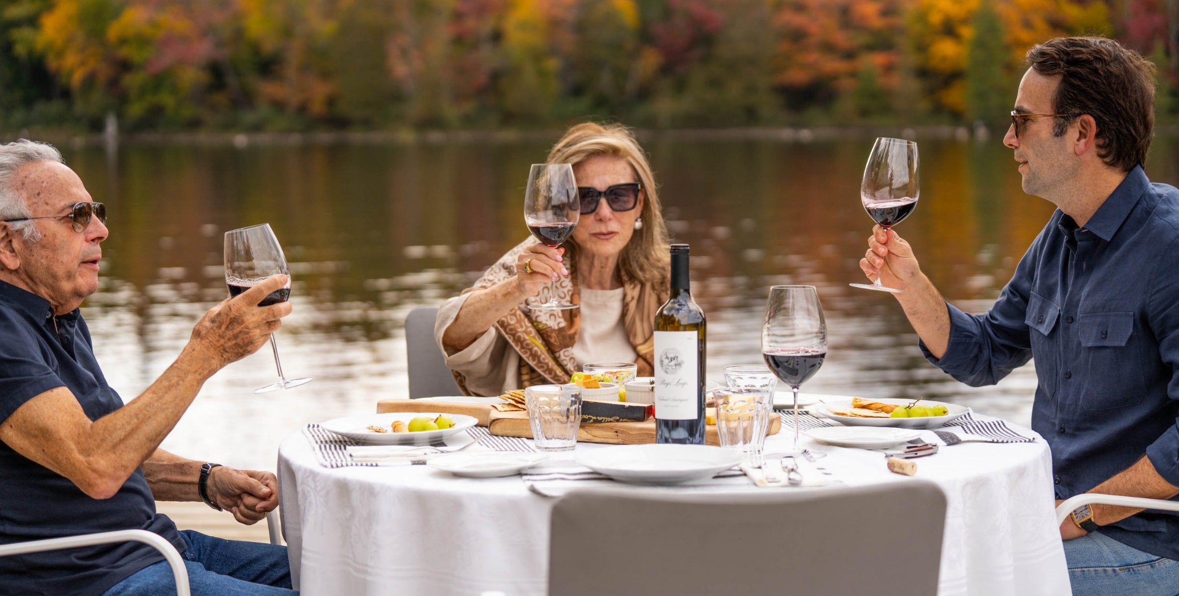 Three people enjoying a meal and wine by a lake.