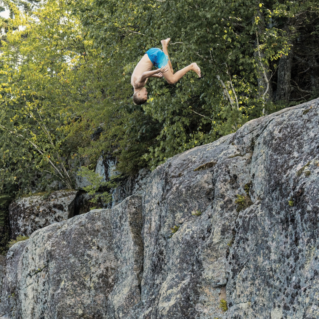 Person diving off a cliff into a body of water with trees in the background