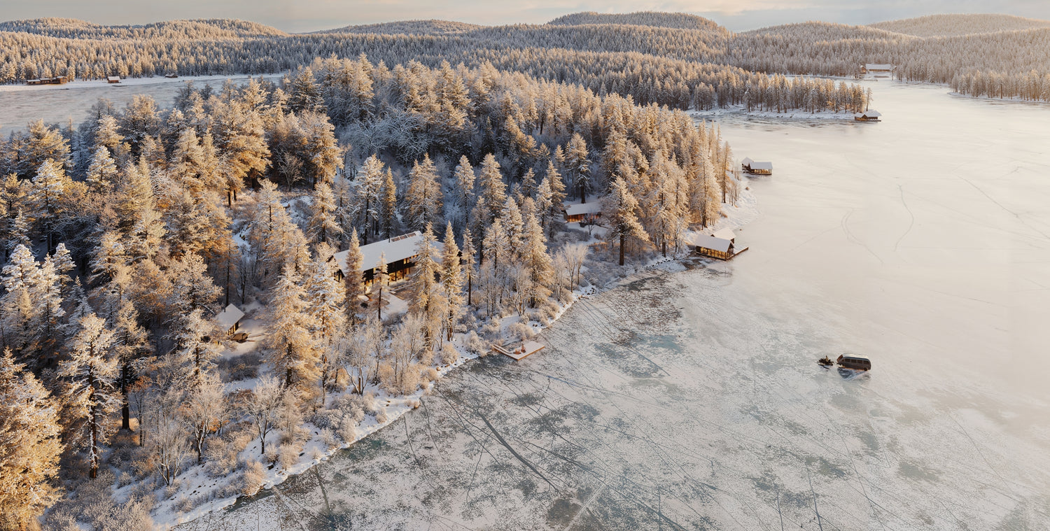 Aerial view of a frozen lake surrounded by snow-covered trees with a cabin.
